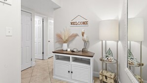 A hallway with a white door, a welcome sign, a vase with pampas grass, a decorative bird, a lamp, and a mirror with a palm tree design.