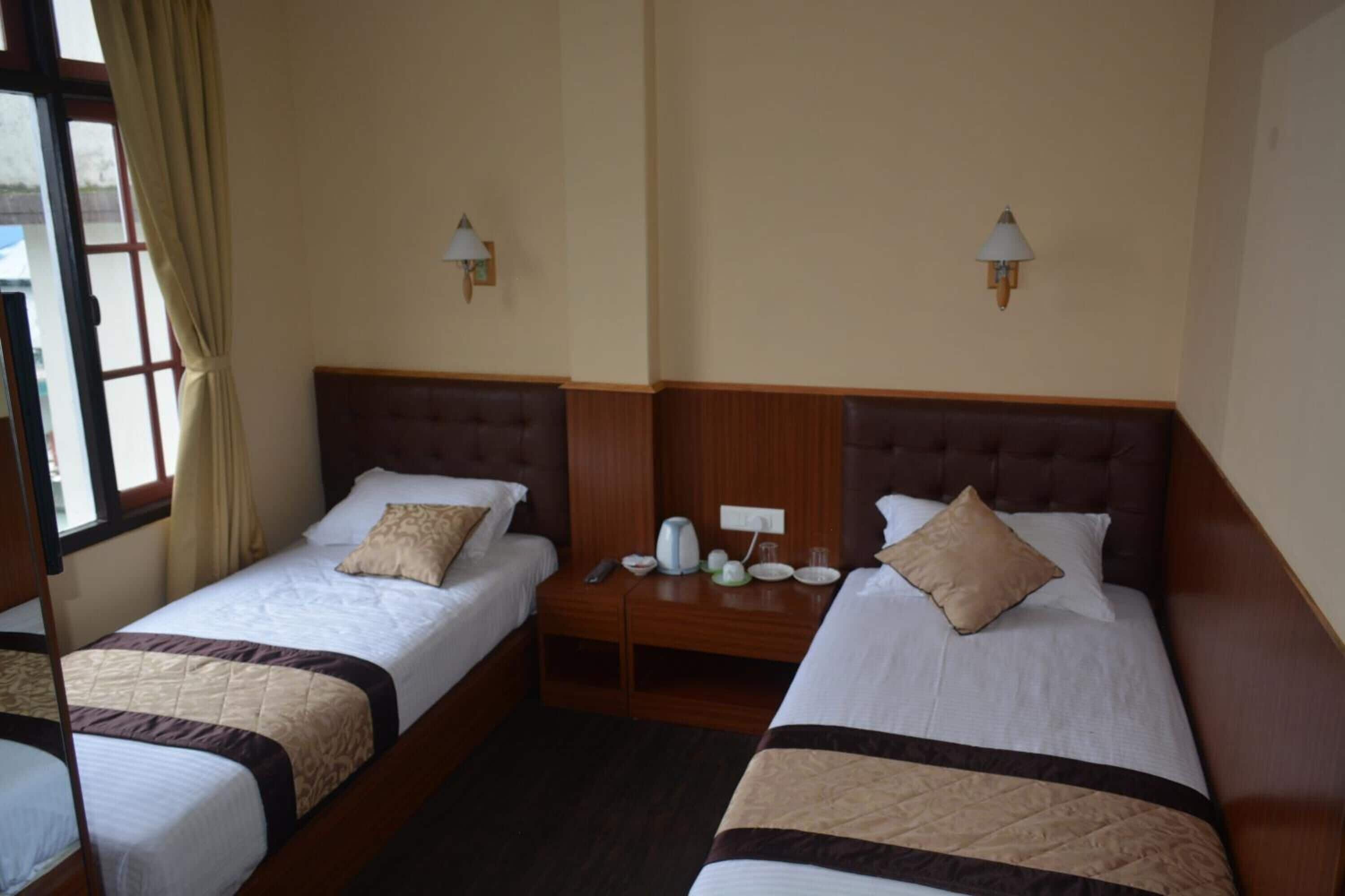 Une chambre d’hôtel avec deux lits simples, des têtes de lit en bois, des tables de chevet avec des lampes et une fenêtre avec des rideaux.