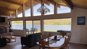 A spacious living area with a fireplace, large windows overlooking a lake, and a dining area with wooden chairs.