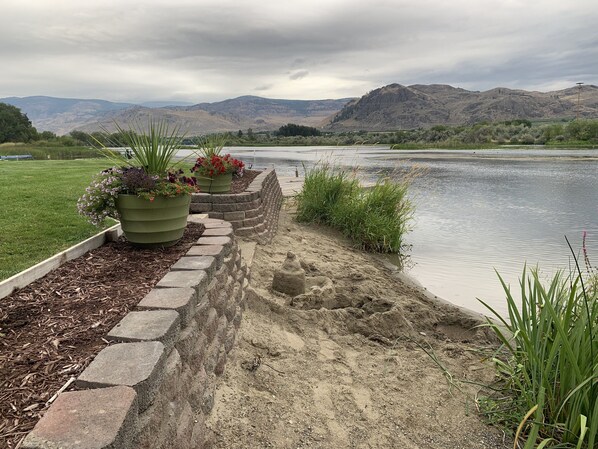 Un paseo a orillas del río con escalones de piedra que conducen a una zona arenosa junto a un río tranquilo, rodeado de vegetación y montañas lejanas.