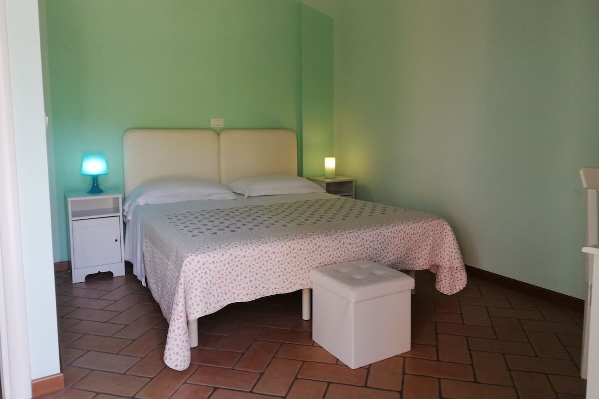 Quarto com cama, duas mesas de cabeceira, um criado-mudo e um pequeno banco branco.