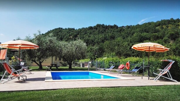 Una piscina con sillones de descanso y sombrillas, rodeada de vegetación y colinas.