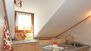 A kitchen with a sink, wooden cabinets, a window with lace curtains, and a fruit-patterned towel. - Casa Gilda (Falcade)