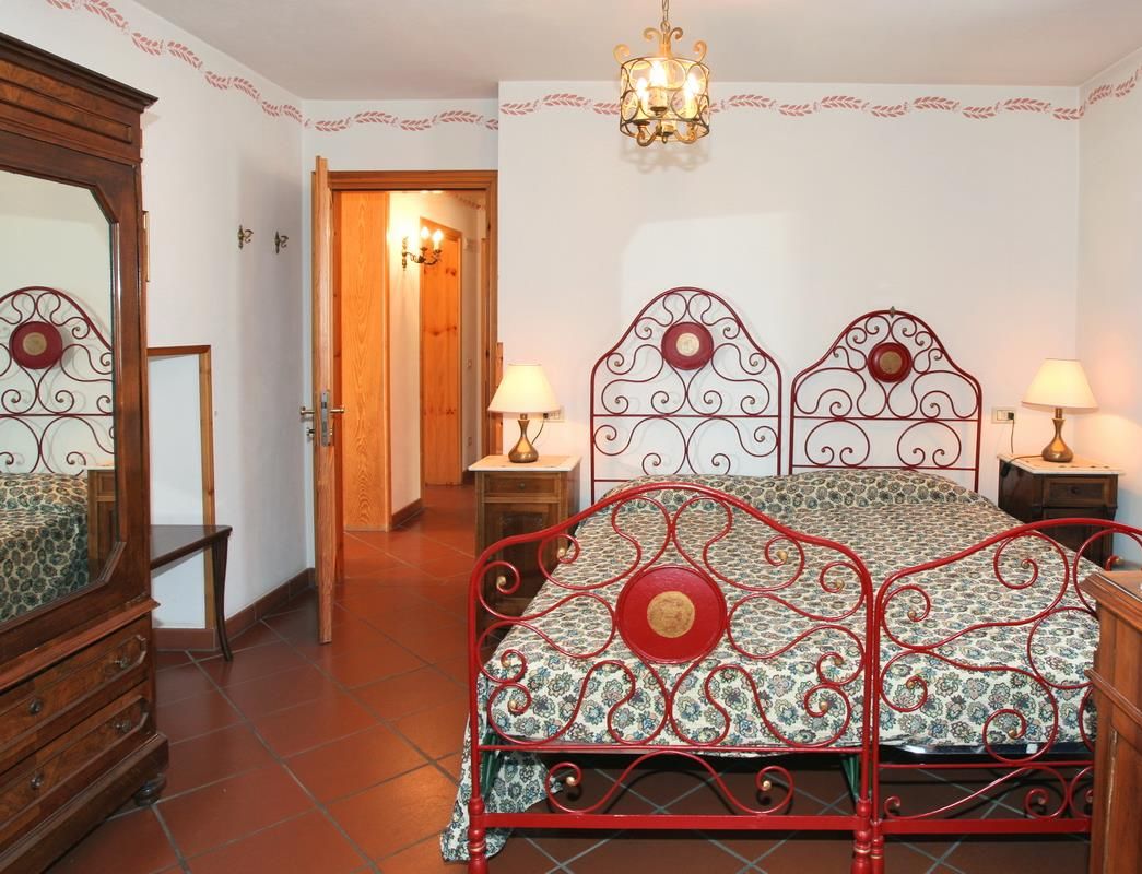 Een slaapkamer met een rode metalen bed, beddengoed met een patroon, twee bedlampjes en een houten dressoir.