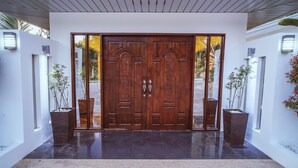 A double wooden door with intricate carvings, flanked by two potted plants, leading to a modern building with a tiled floor and a white exterior. - Quest Villa (Panglao)