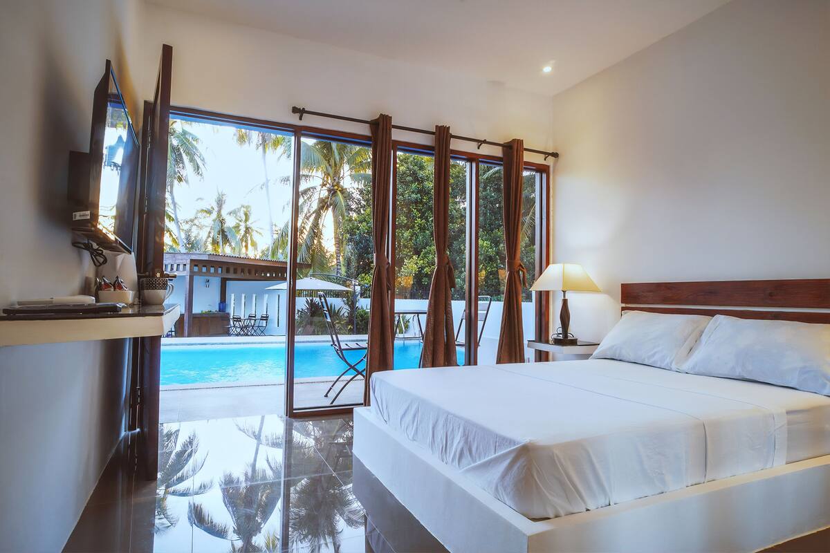 A hotel room with a large bed, a TV, and a view of a swimming pool and palm trees.