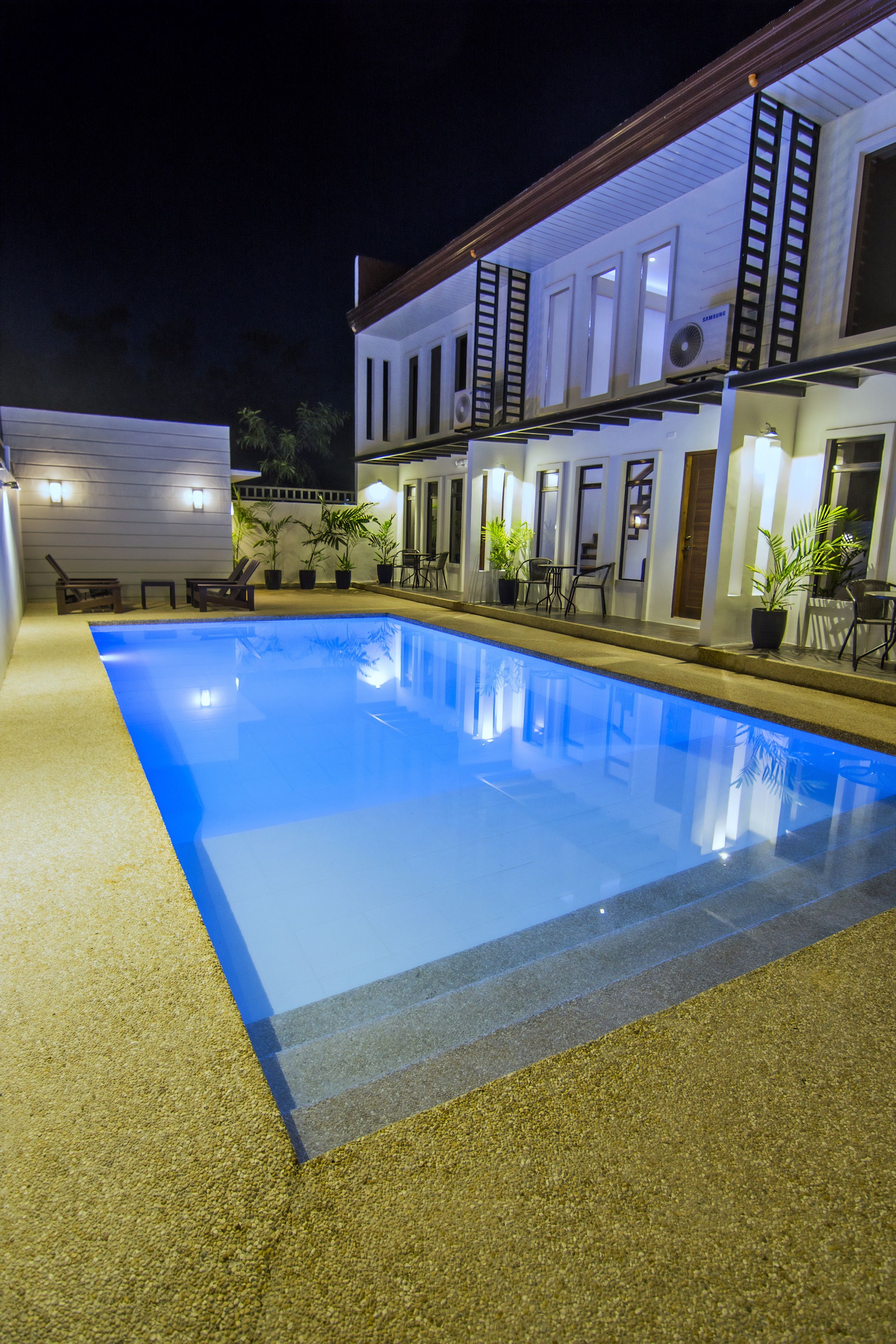 A swimming pool with a tiled deck, adjacent to a building with multiple doors and windows, and outdoor seating.