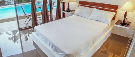 Una cama grande con ropa blanca, dos lámparas de noche y vista a una piscina a través de un ventanal.