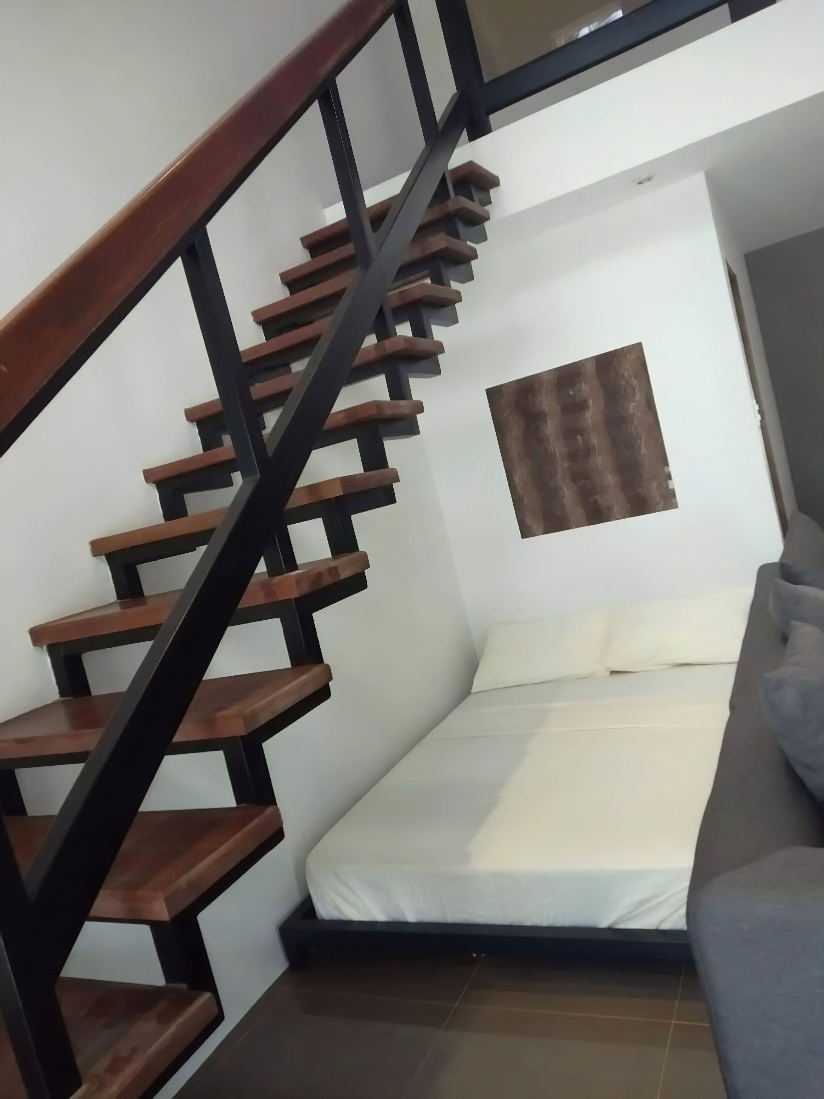 A bedroom with a wooden staircase, a bed with white bedding, and a small window with a brown curtain.