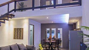 A modern living room with a sofa, dining table, and chairs, a staircase leading to an upper level, and a refrigerator. - Quest Villa (Panglao)