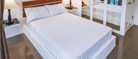 Una cama grande con ropa de cama blanca, dos mesitas de noche con lámparas, un cabecero de madera y un espejo grande que refleja una zona de piscina.