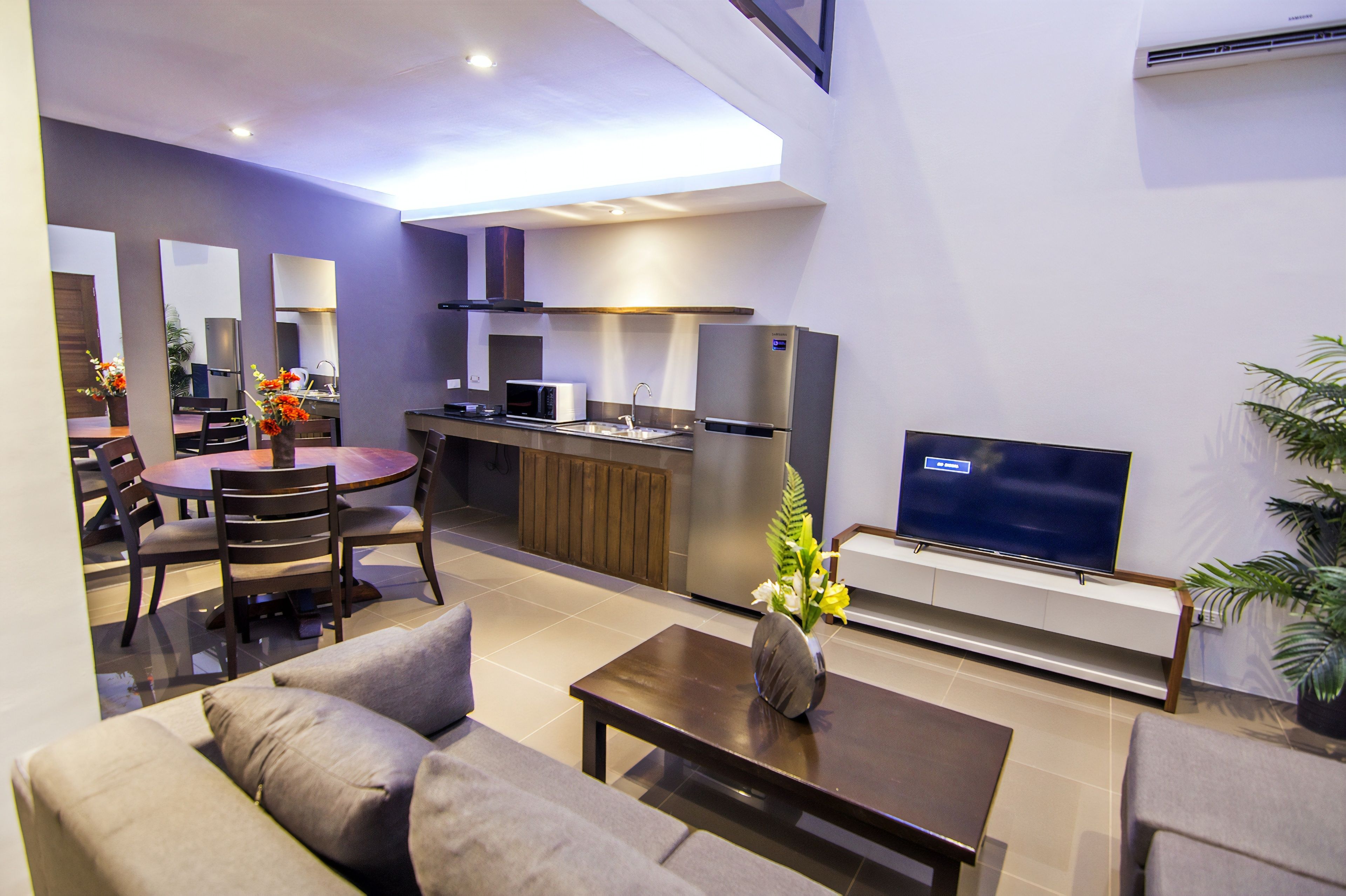 A modern living room with a sofa, coffee table, and a TV. There is a kitchen area with a microwave, refrigerator, and sink.