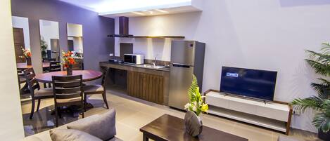 A modern living room with a sofa, coffee table, and a TV. There is a kitchen area with a microwave, refrigerator, and sink.