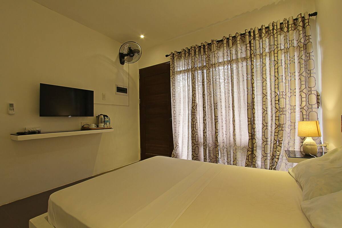 A hotel room with a bed, a TV mounted on the wall, a bedside table with a lamp, and a curtain with a geometric pattern.