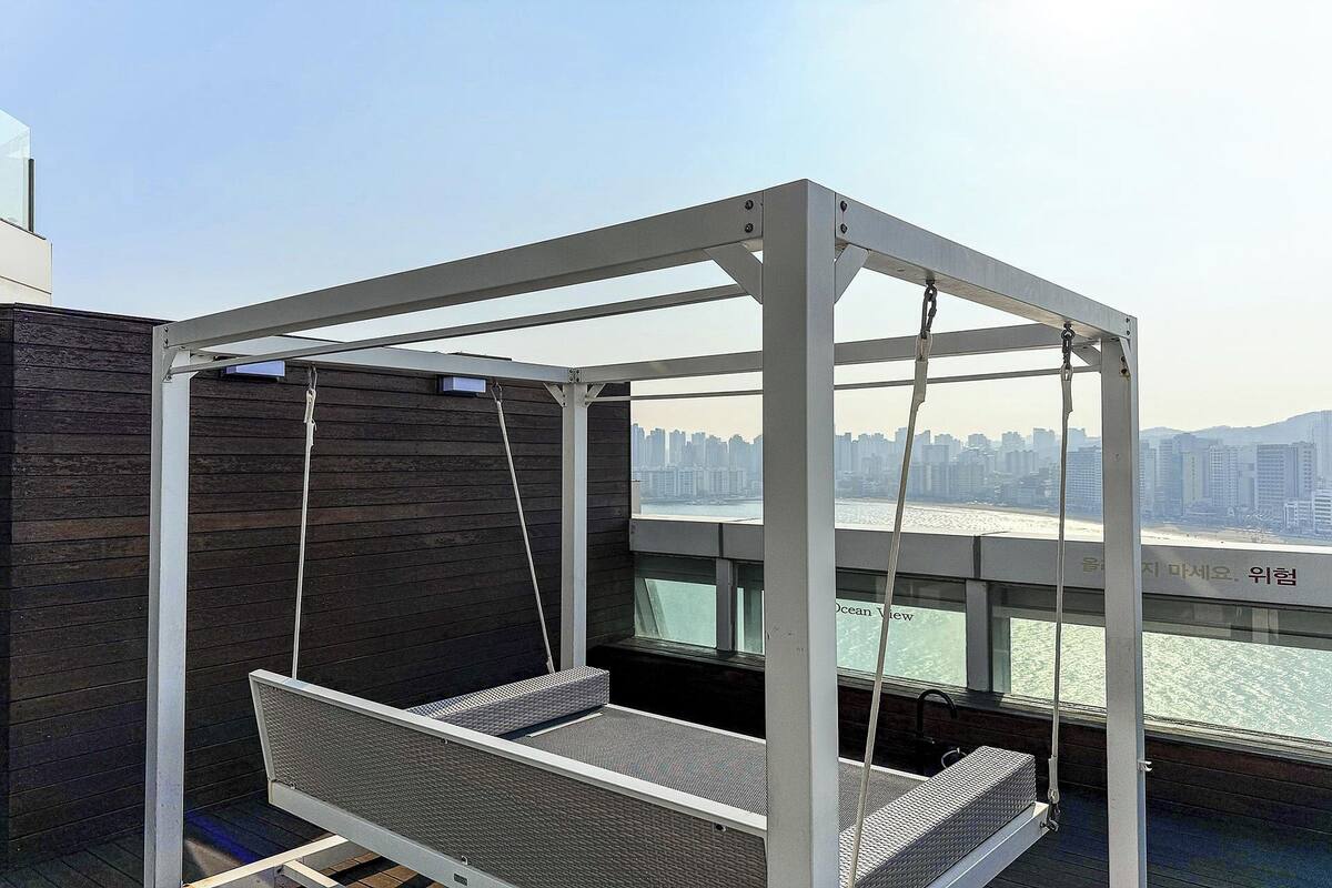 A white swing chair with a metal frame, a window with a view of a city skyline, and a wooden wall.