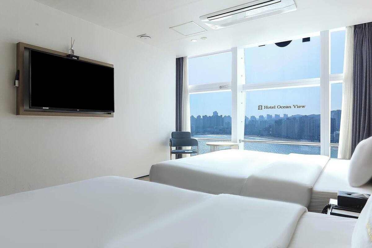 A hotel room with two beds, a TV, and a large window with a city view.