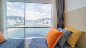A room with a large window offering a view of a city skyline and a beach. There are two blue sofas with orange and yellow cushions.