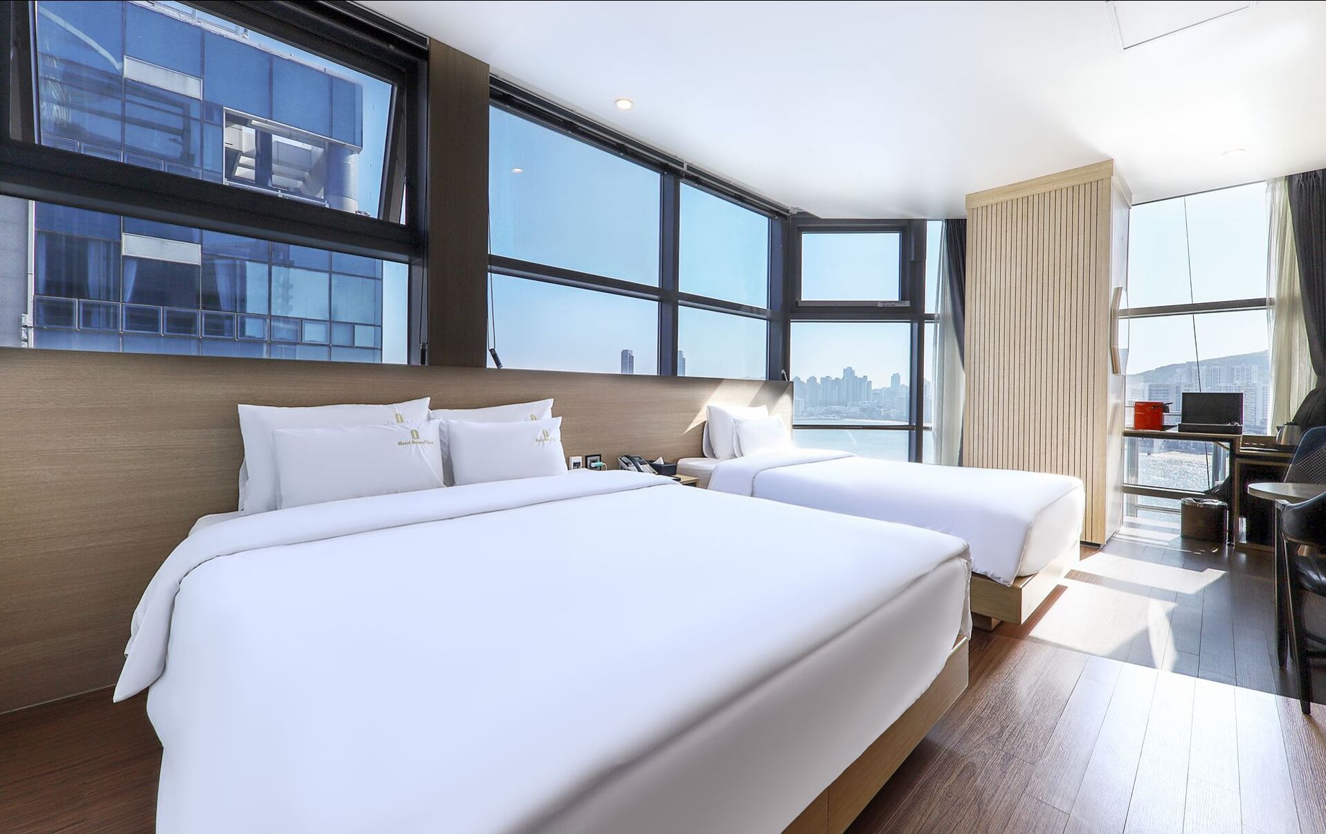 A hotel room with two beds, large windows, a city view, and a desk with a laptop.
