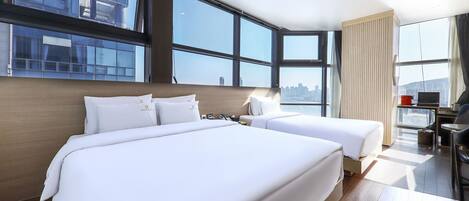 A hotel room with two beds, large windows, a city view, and a desk with a laptop.