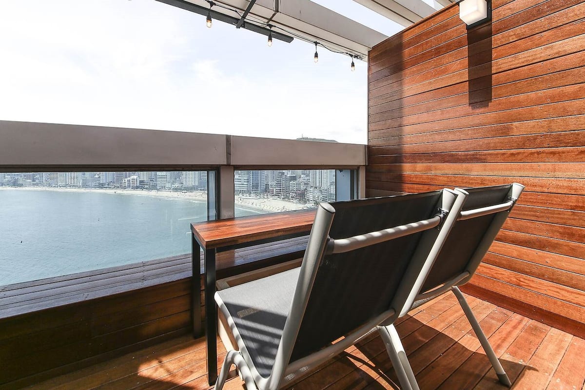 A balcony with a wooden floor, a table, and two chairs. A cityscape and beach are visible through the window.