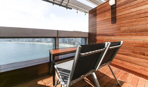 A balcony with a wooden floor, a table, and two chairs. A cityscape and beach are visible through the window. - Gwanganli Hotel Ocean View (Busan)