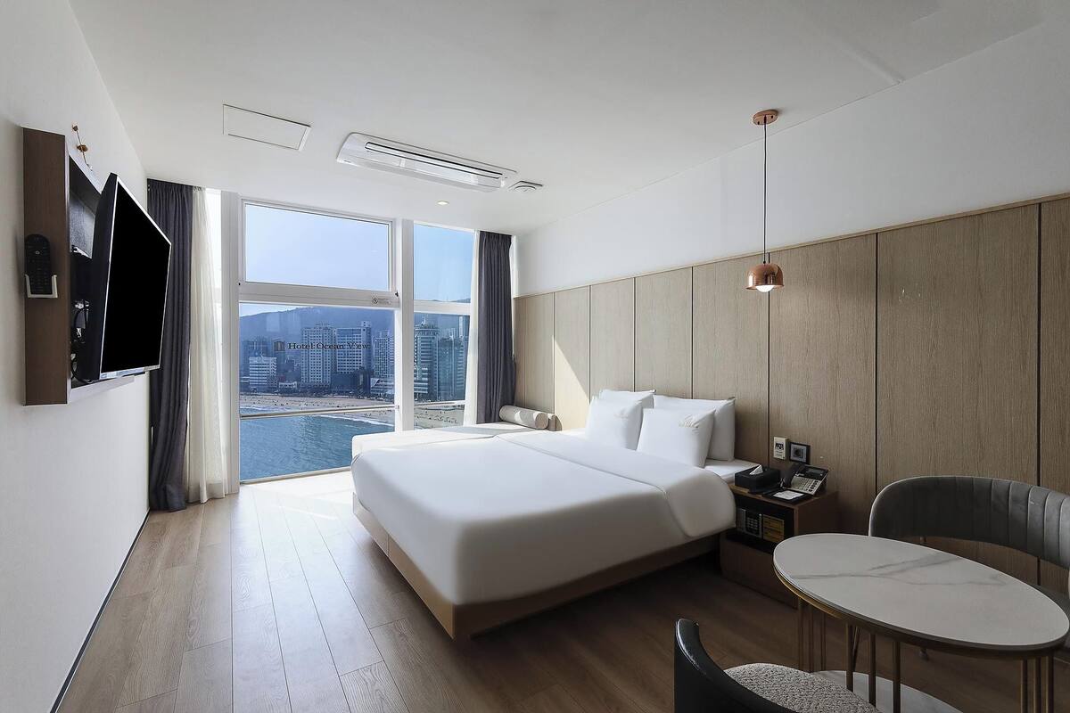A hotel room with a large bed, a desk with a chair, a TV, and a view of the cityscape.
