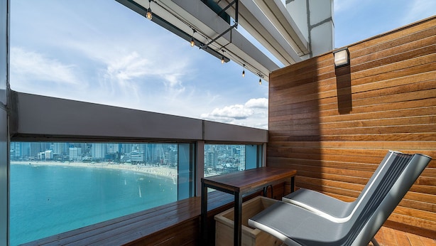 A balcony with a wooden floor, a metal table, and a metal chair. A wooden wall and a window with a view of a beach and cityscape are visible.