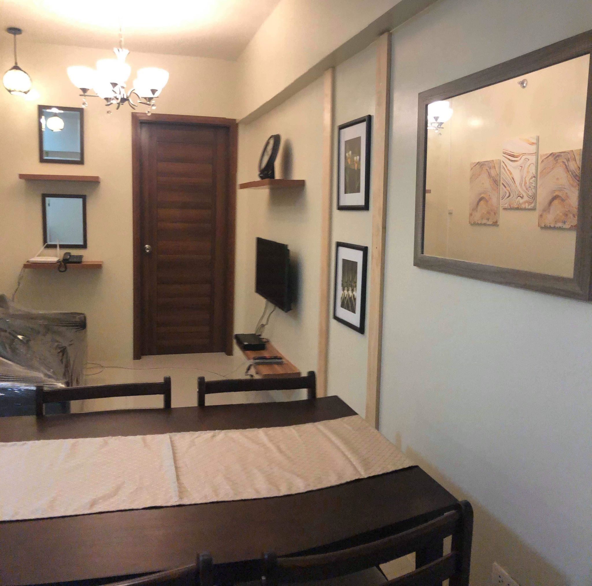 Une pièce avec une porte en bois, un bureau avec un ordinateur, une chaise, un lit recouvert d’une couverture blanche, et un mur orné de tableaux encadrés et d’une peinture.
