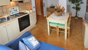 A cozy kitchen with a stove, sink, and cabinets. There's a dining table with chairs and a blue sofa with cushions. A potted plant and a television are also visible. - Casa Quercia (Falcade)