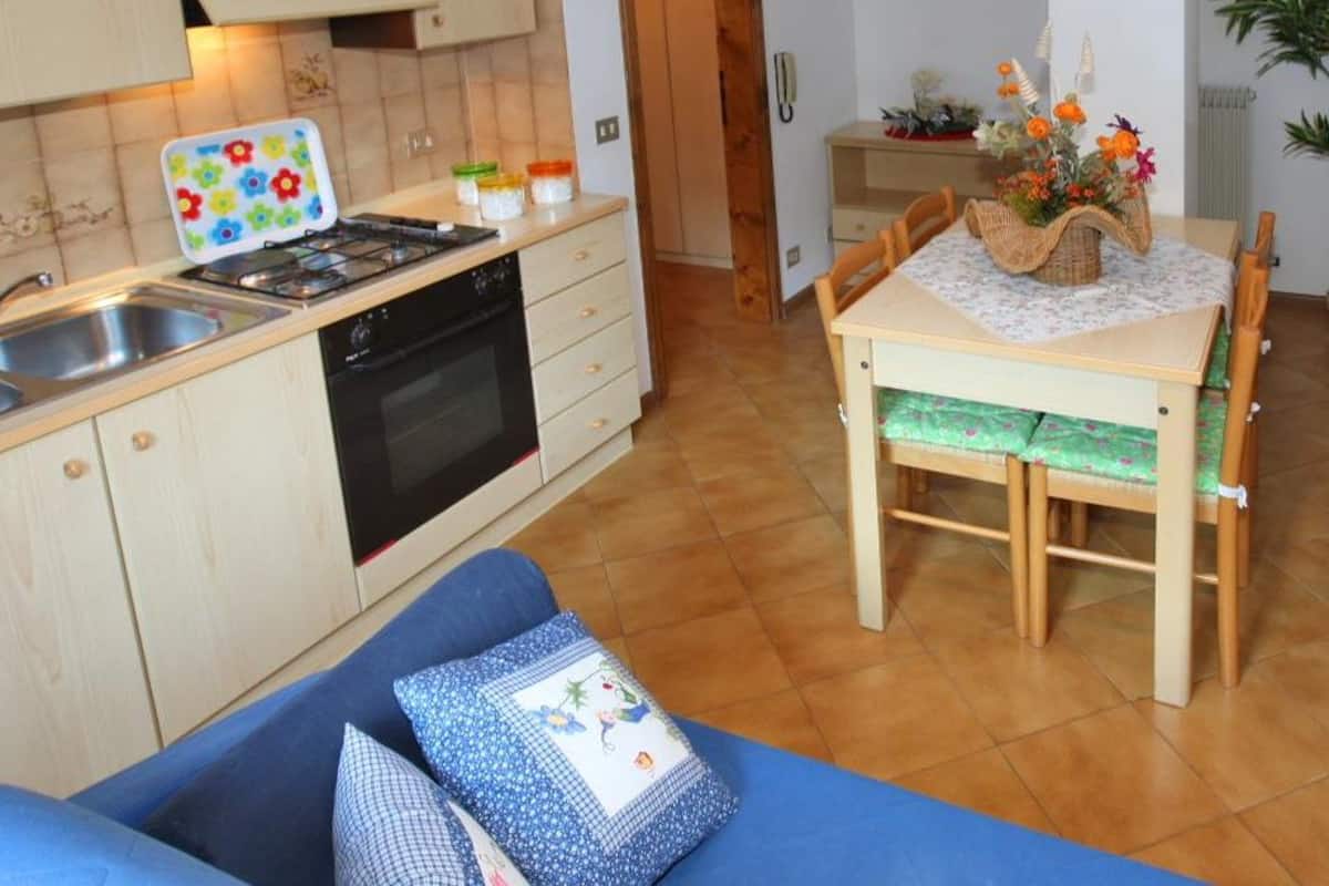 Une cuisine chaleureuse avec un poêle, un évier et des placards. On y trouve une table à manger avec des chaises, ainsi qu’un canapé bleu avec des coussins. Une plante en pot et un téléviseur sont également visibles.