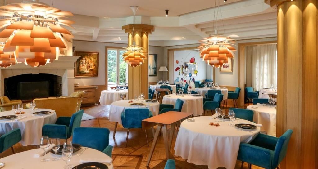 Un espace repas avec des tables rondes, des chaises bleues et des lampes suspendues orange.