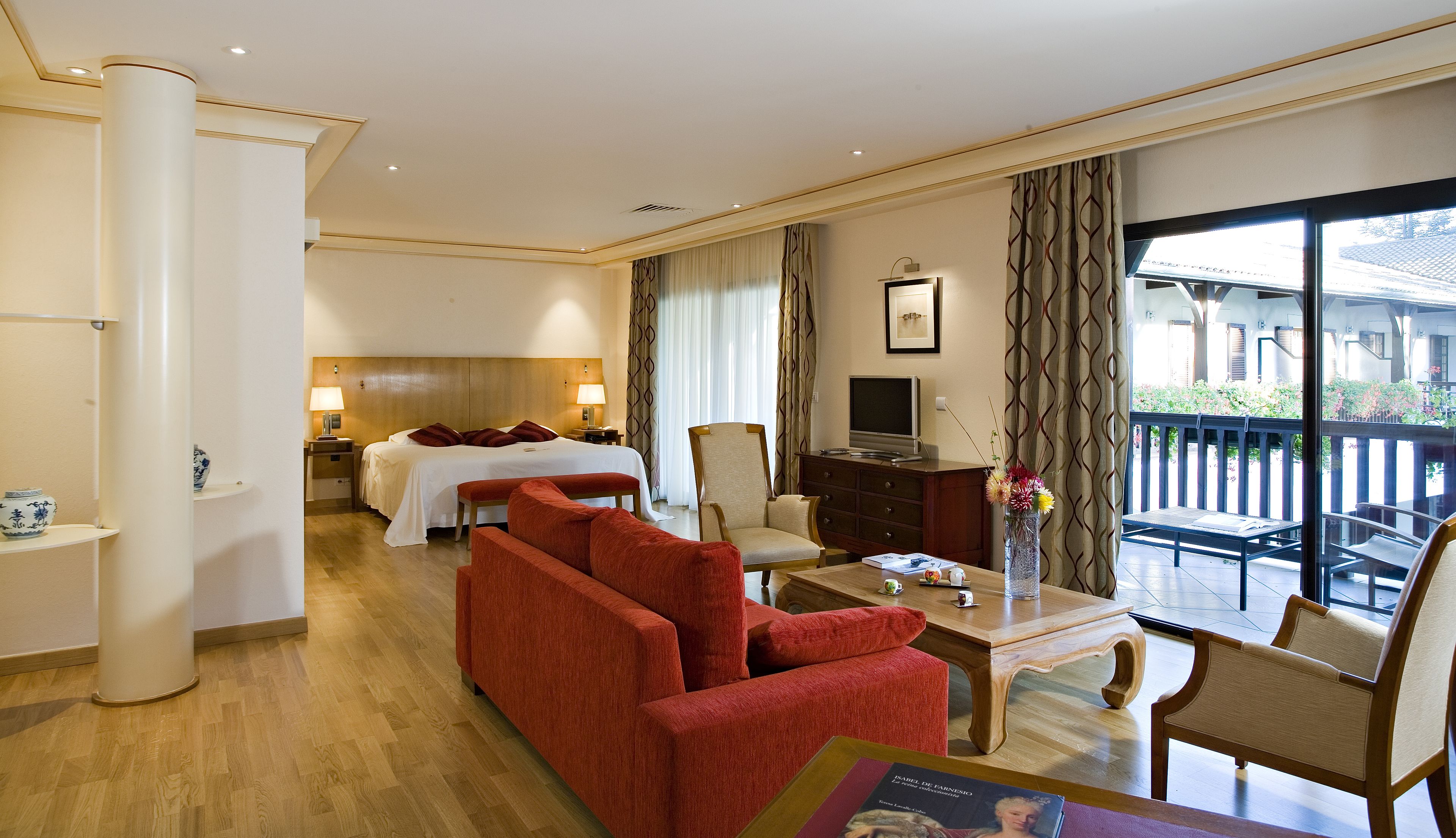 A hotel room with a bed, a red sofa, a wooden coffee table, a TV, and a balcony with a view.