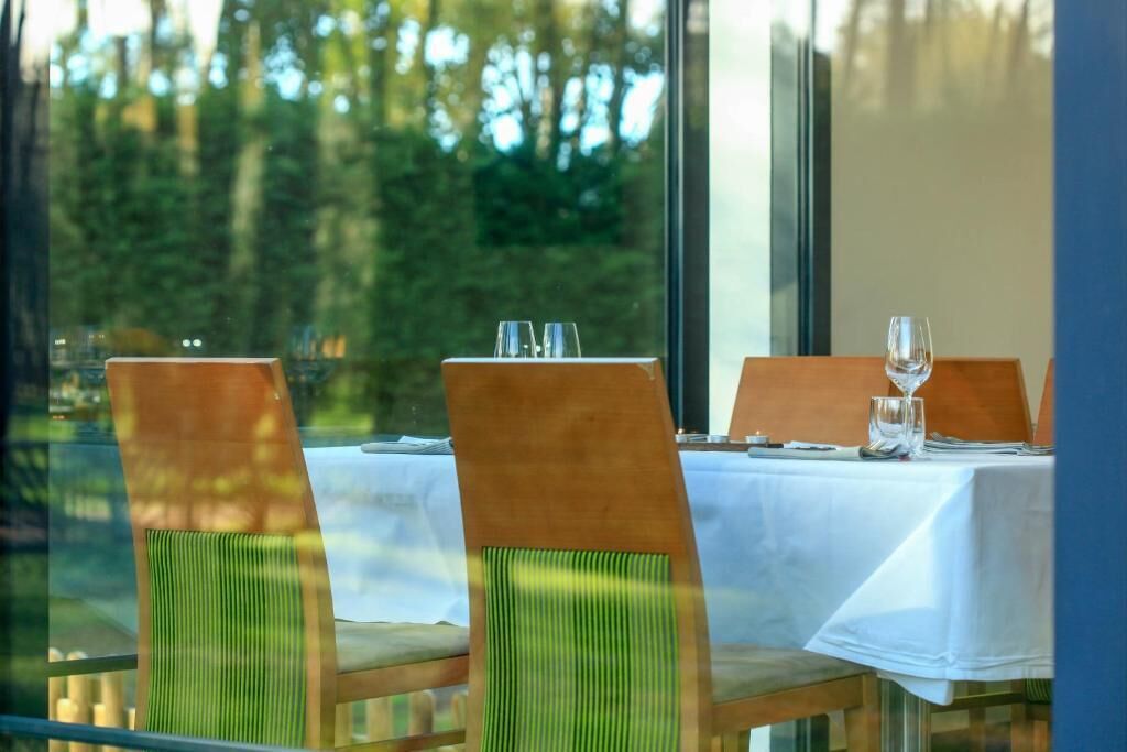 A dining area with a table set for two, chairs with green cushions, and a view of trees through large windows.
