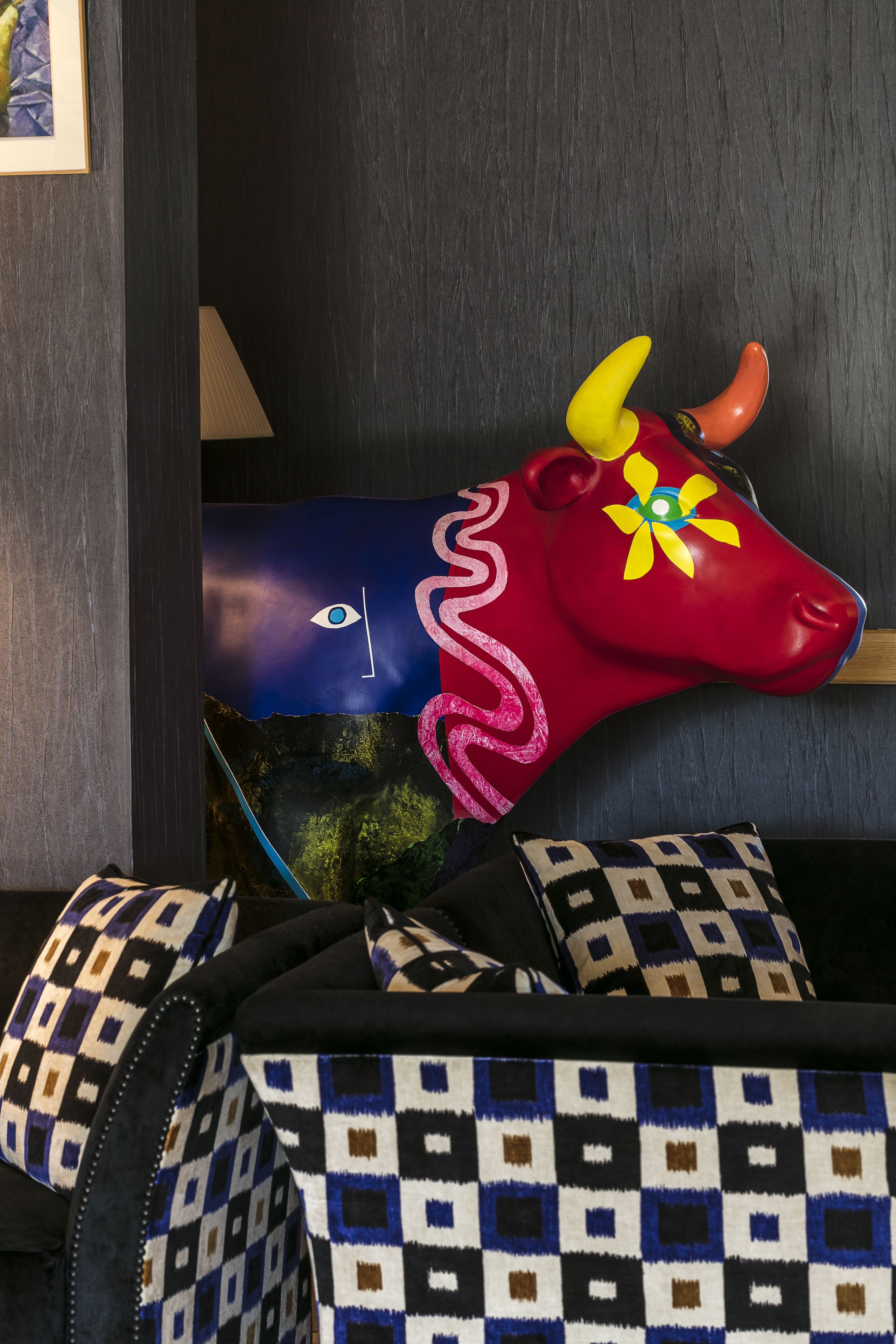 A room with a colorful cow decoration, patterned pillows, and a dark wooden wall.