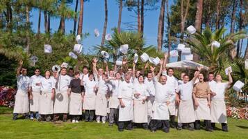 A group of people in white chef uniforms standing in a grassy outdoor area with trees in the background.