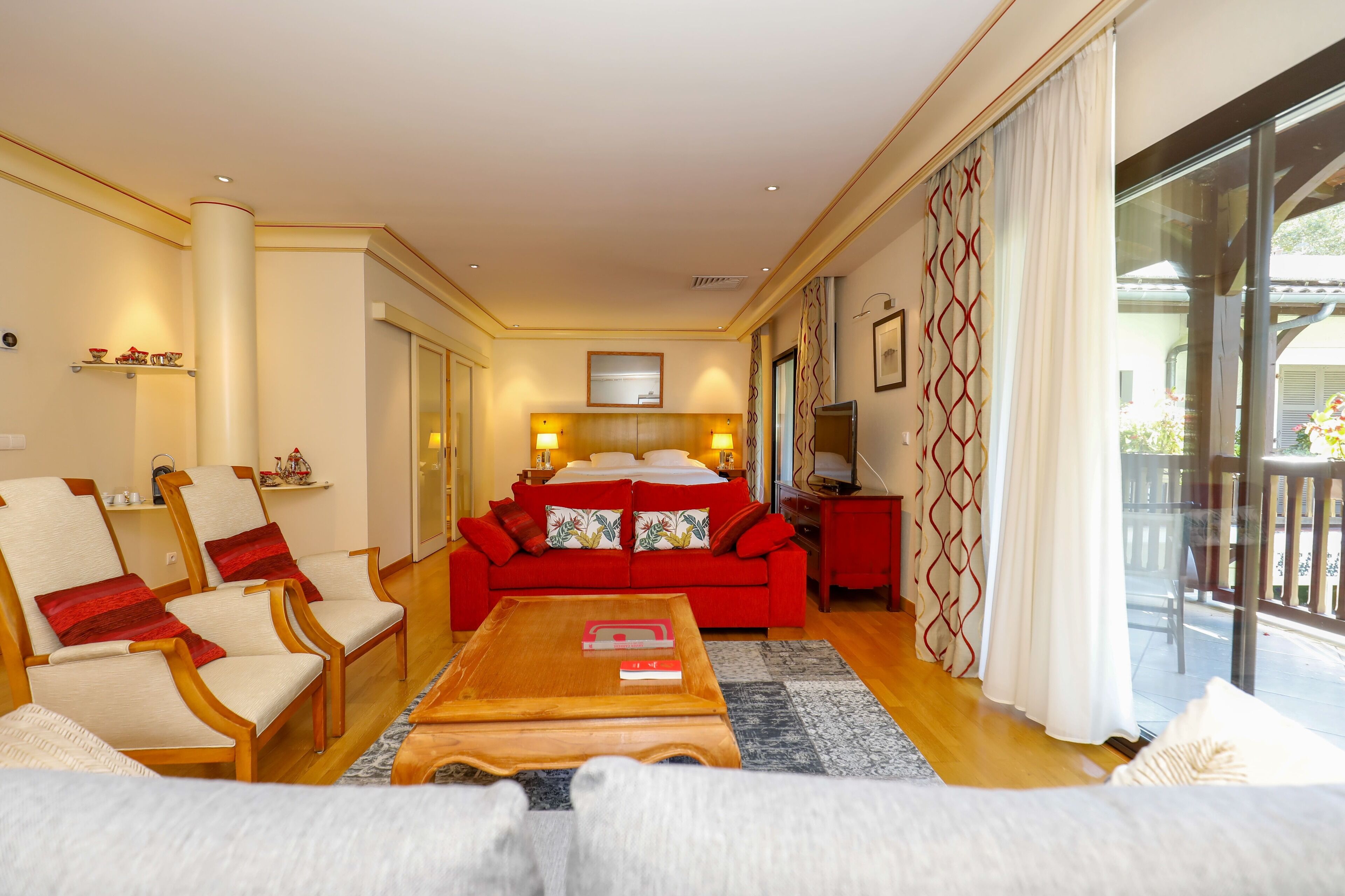 Una habitación de hotel con un sofá rojo, sillas de madera, una mesa de centro de madera y un ventanal con cortinas.
