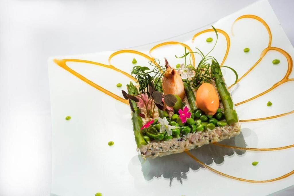 Un plato cuadrado con una variedad de ingredientes y adornos, presentado sobre un plato blanco con un diseño de línea amarilla decorativa.