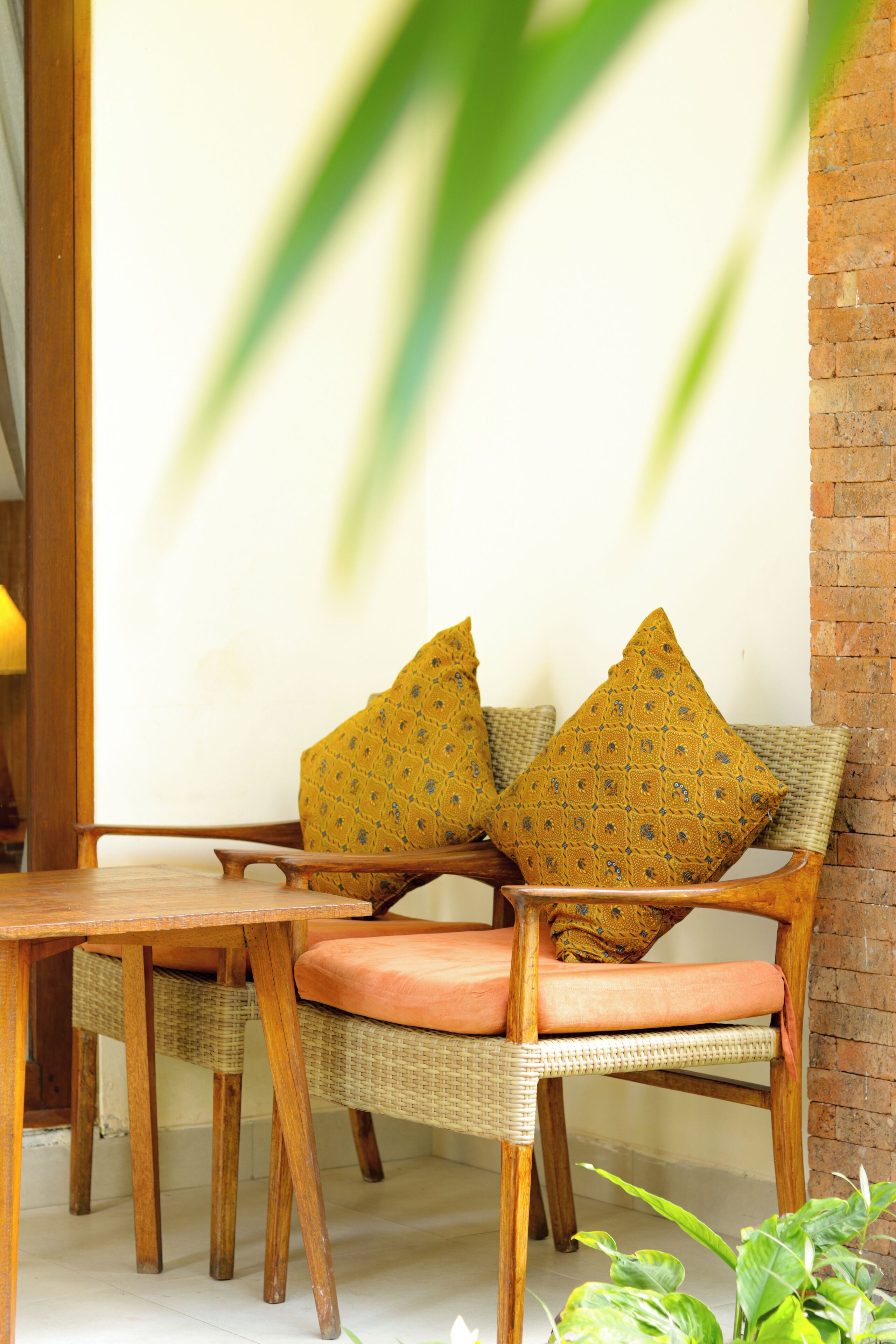 A wicker chair with orange cushions and patterned pillows, a wooden table, and a brick wall.