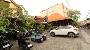 A street with parked scooters and a white car, a building with a tiled roof, and a restaurant with outdoor seating. - Mina Pelasa Hotel and Resto (Legian)