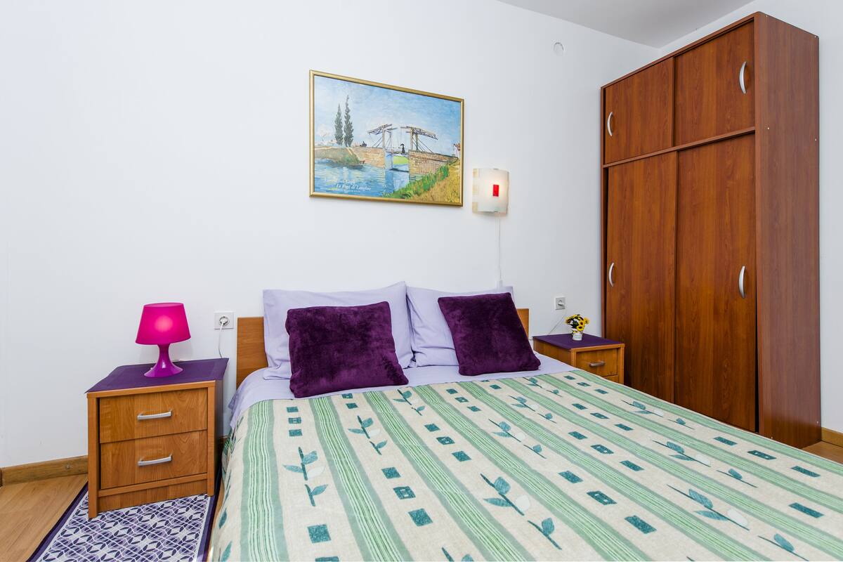 A bedroom with a bed, a nightstand, a lamp, a wardrobe, and a painting on the wall.