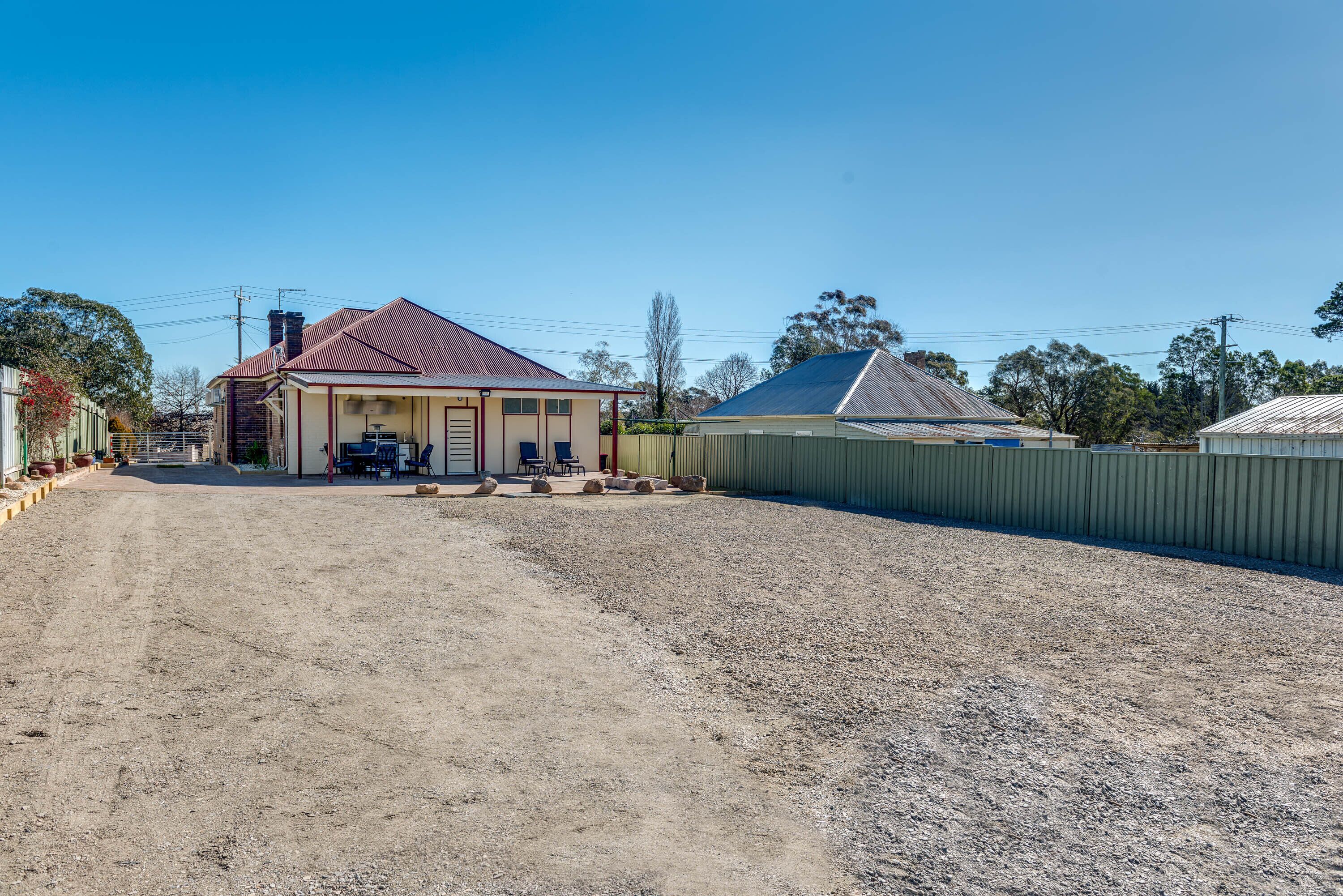 A property with a dirt driveway leading to a house with a red roof and a fenced yard.