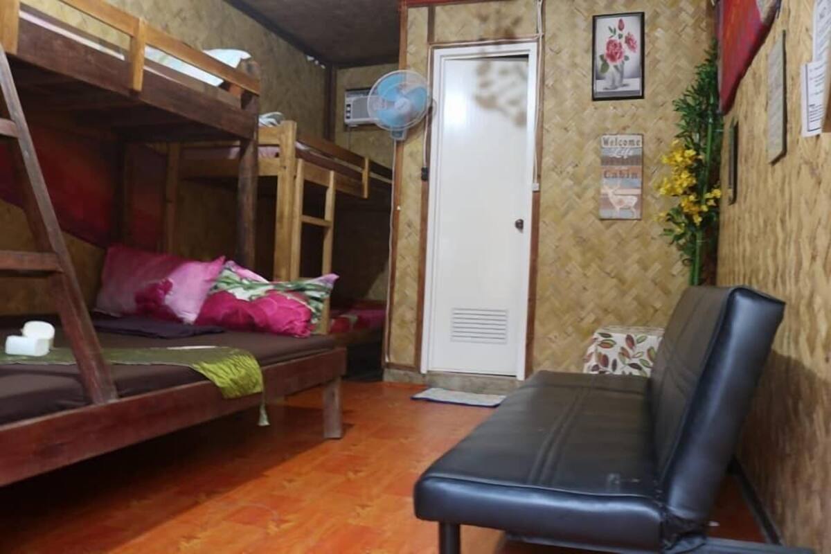 Uma cama beliche com escada de madeira, um sofá preto e uma porta branca com um ventilador.