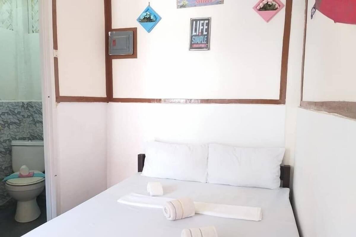 Quarto de hotel com cama, roupa de cama branca, uma pequena mesa de cabeceira e uma parede com um detector de fumaça e um porta-retrato.