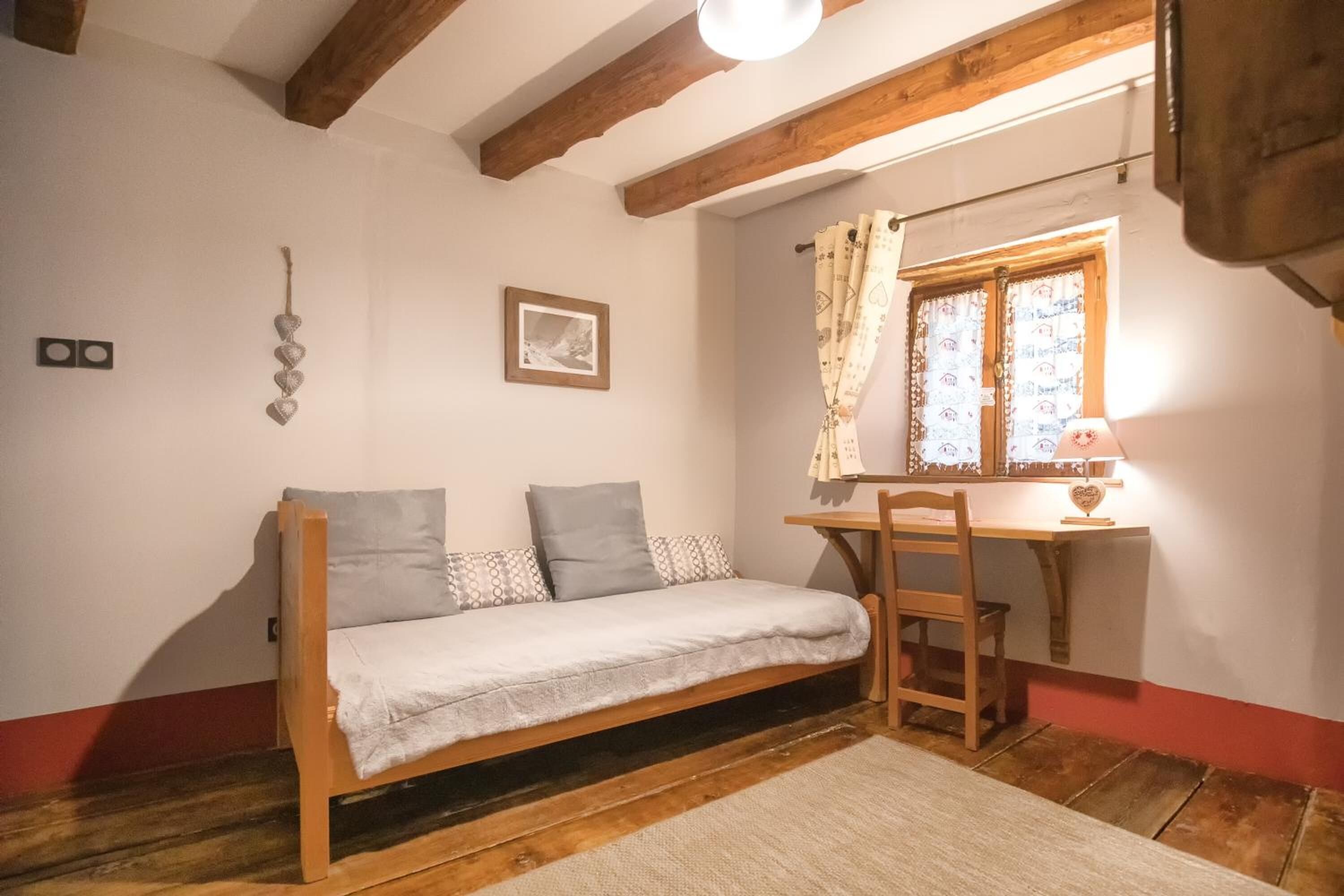 Una stanza piccola e accogliente con un letto di legno, una scrivania con una sedia, una finestra con tende e un lampadario a parete.