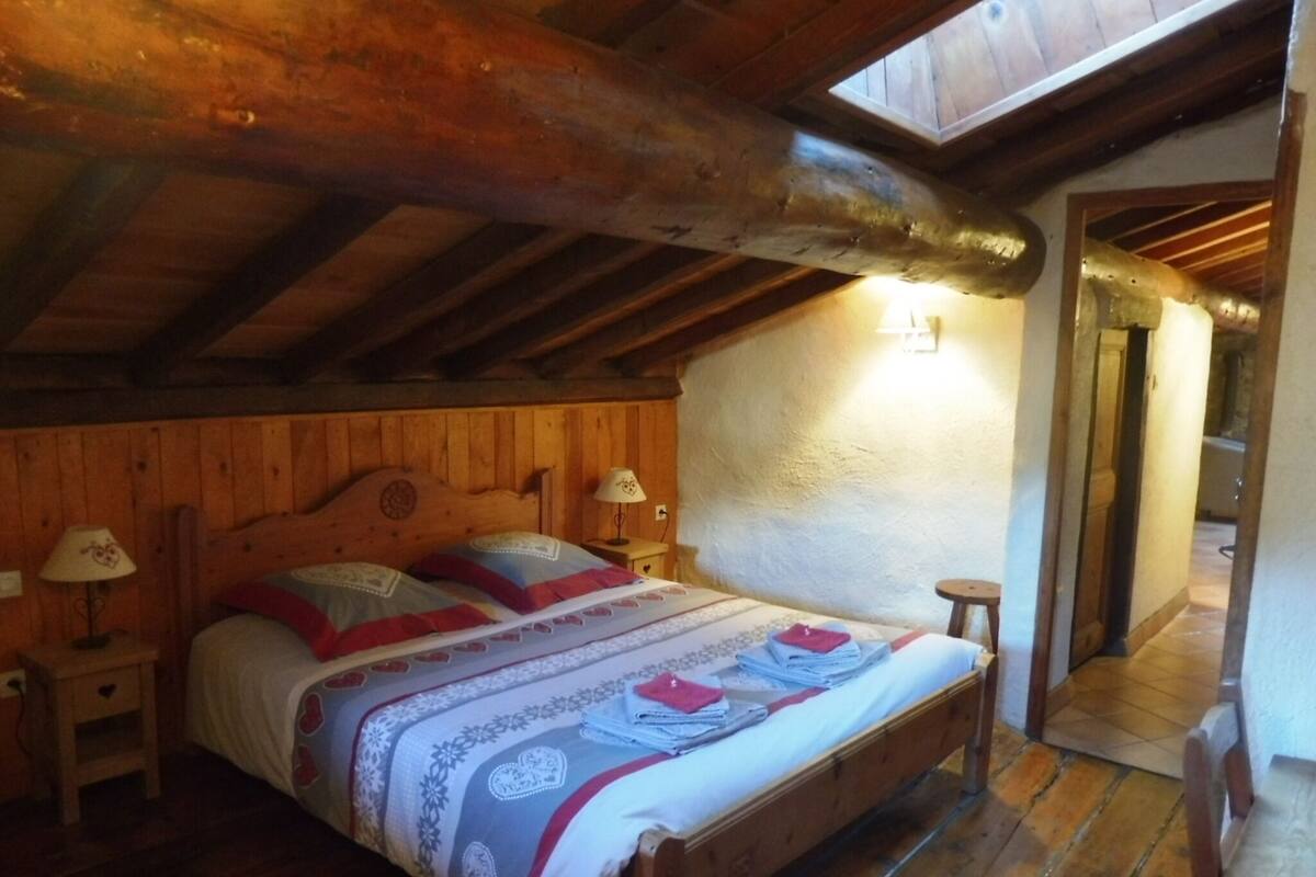 Un dormitorio acogedor con techo de madera, una cama con colcha estampada, dos mesitas de noche con lámparas y un taburete pequeño.