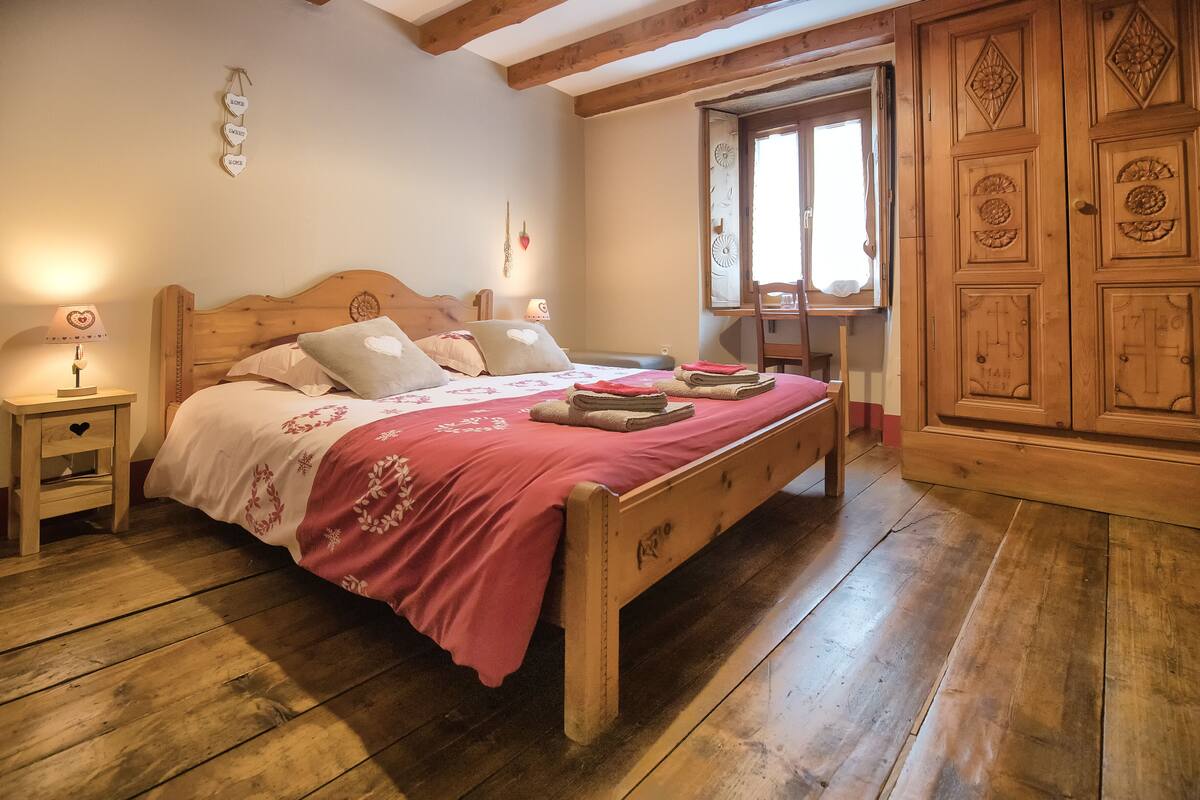 Una cama de madera con una colcha roja, mesitas de noche de madera y una ventana con cortinas.