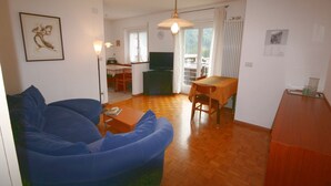 A living room with a blue sofa, a wooden dining table, and a TV in the background. - Casa Angelo (Falcade)