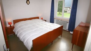 A bedroom with a wooden bed, two bedside lamps, a radiator, a window with blue curtains, and a view of a building outside. - Casa Angelo (Falcade)