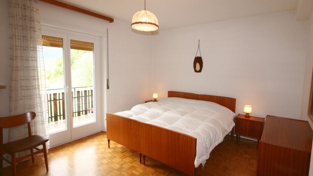 A bedroom with a wooden bed, two bedside tables with lamps, a wooden chair, a wooden desk, and a large window with a view of the outdoors. - Casa Angelo (Falcade)