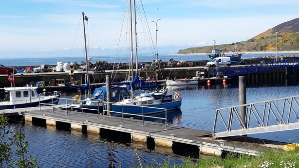 A harbor with boats docked, a pier, and a hilly landscape in the background.